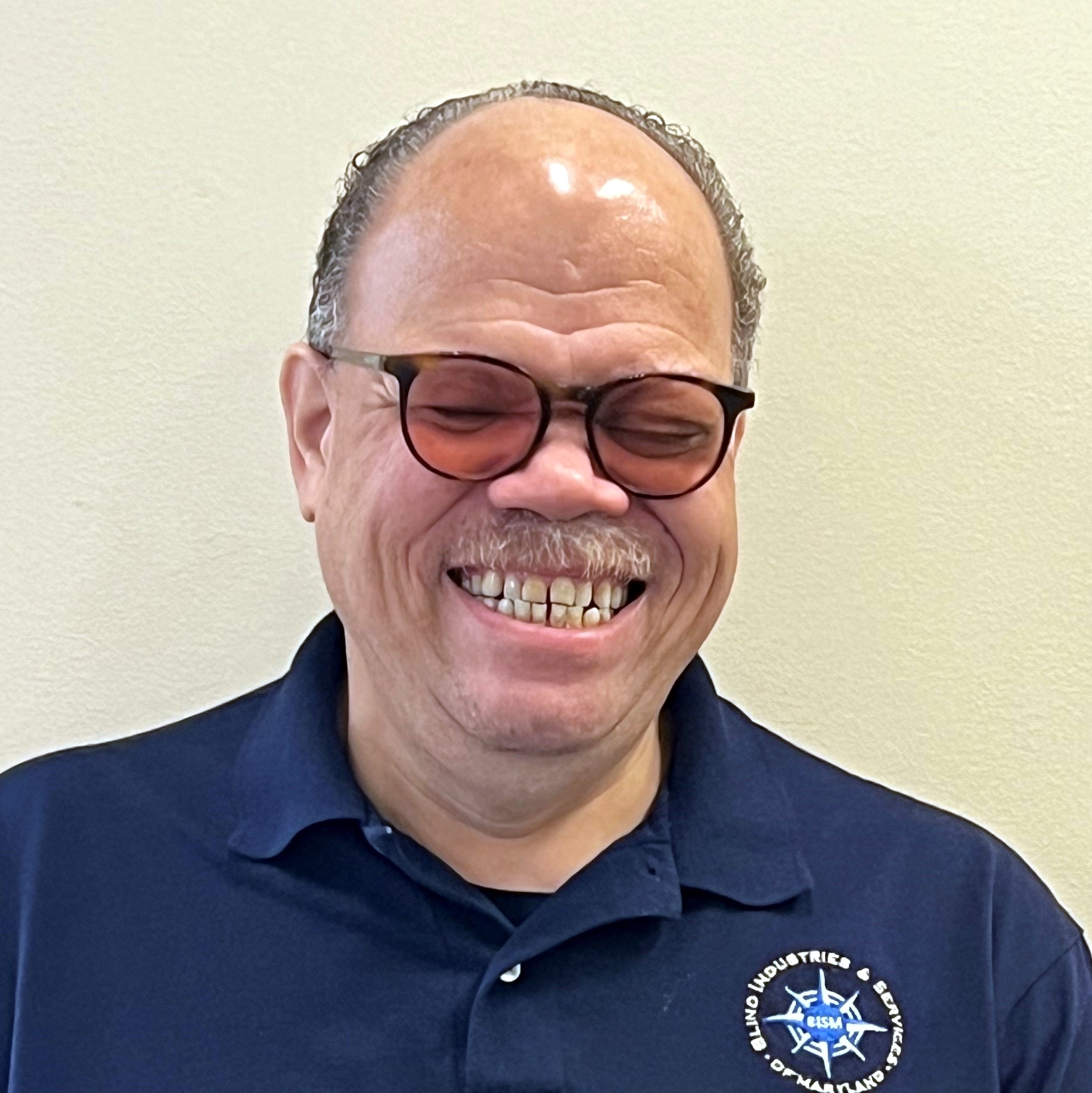Photo of Kevin standing in front of a white wall. He wears a pair of glasses, long-sleeve Blue BISM polo, and a fantastic smile.